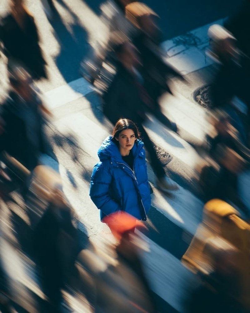 A person in a blue jacket standing calm and in focus on a city crosswalk while the crowd around them blurs past, representing someone who chooses to stand out and build their own way